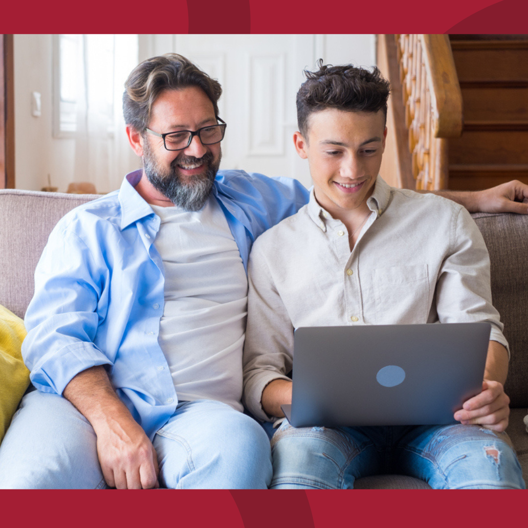 A father and son sitting on a couch together, looking at a laptop.